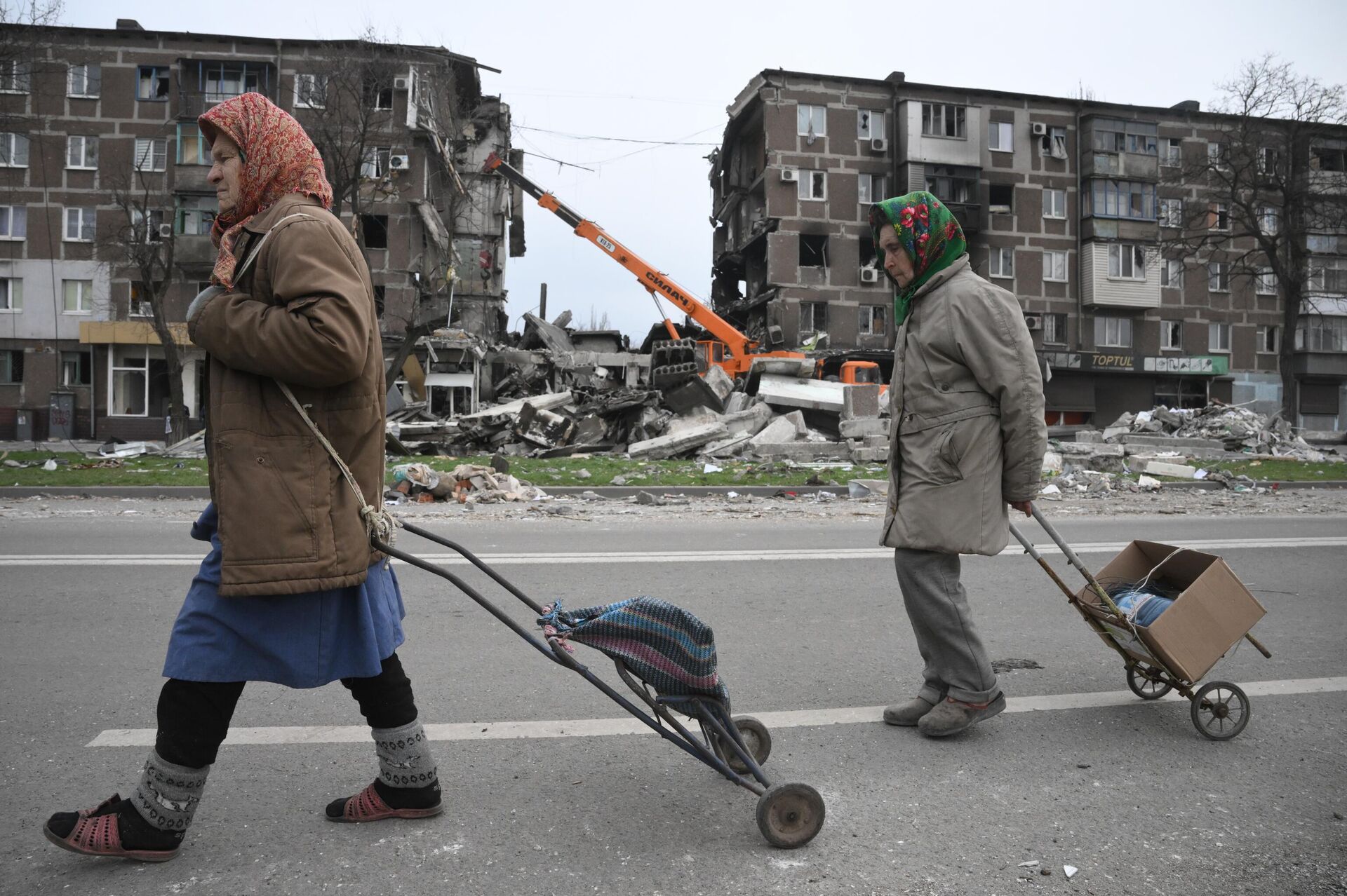 Жители Мариуполя идут по улице города. На втором плане: сотрудники МЧС ДНР разбирают завалы у разрушенных домов - Sputnik Latvija, 1920, 14.04.2022