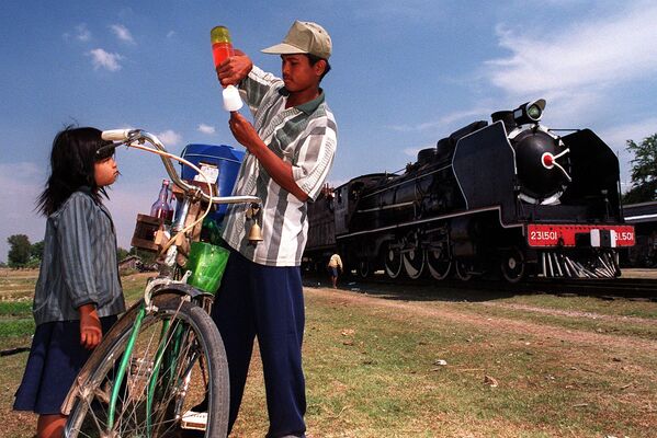Камбоджиец готовит мороженое для маленькой девочки на фоне прибытия старого паровоза &quot;Пасифик 231&quot; на небольшой станции в 50 километрах к югу от Пномпеня, 6 февраля 2000 года.  - Sputnik Латвия